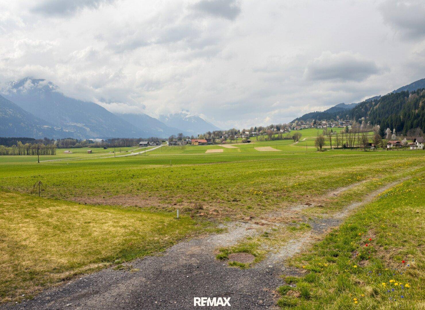 Panoramablick über weite grüne Felder auf ein entferntes Dorf und bewaldete Berge unter einem bewölkten Himmel.