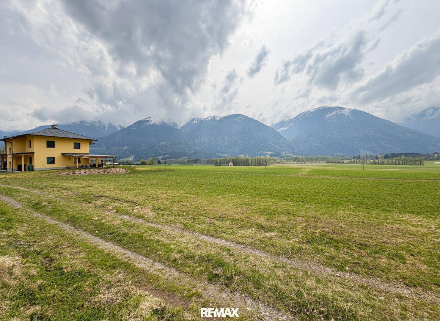 Ein gelbes Haus am Rande weitläufiger grüner Felder mit schneebedeckten Bergen im Hintergrund unter bewölktem Himmel.