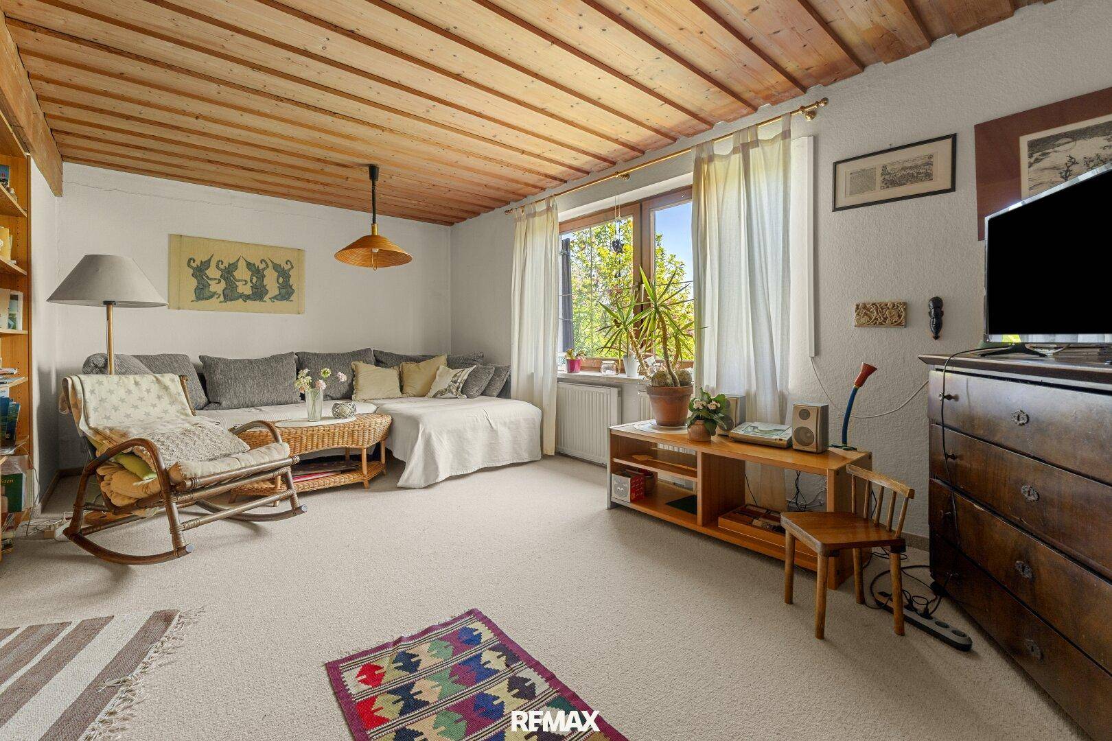 Gemütliches Wohnzimmer mit Teppichboden, Holzbalkendecke und Blick in den Garten.