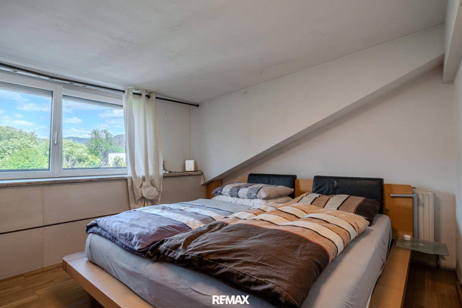 Helles Schlafzimmer mit Holzboden, großem Fenster und Blick ins Grüne.