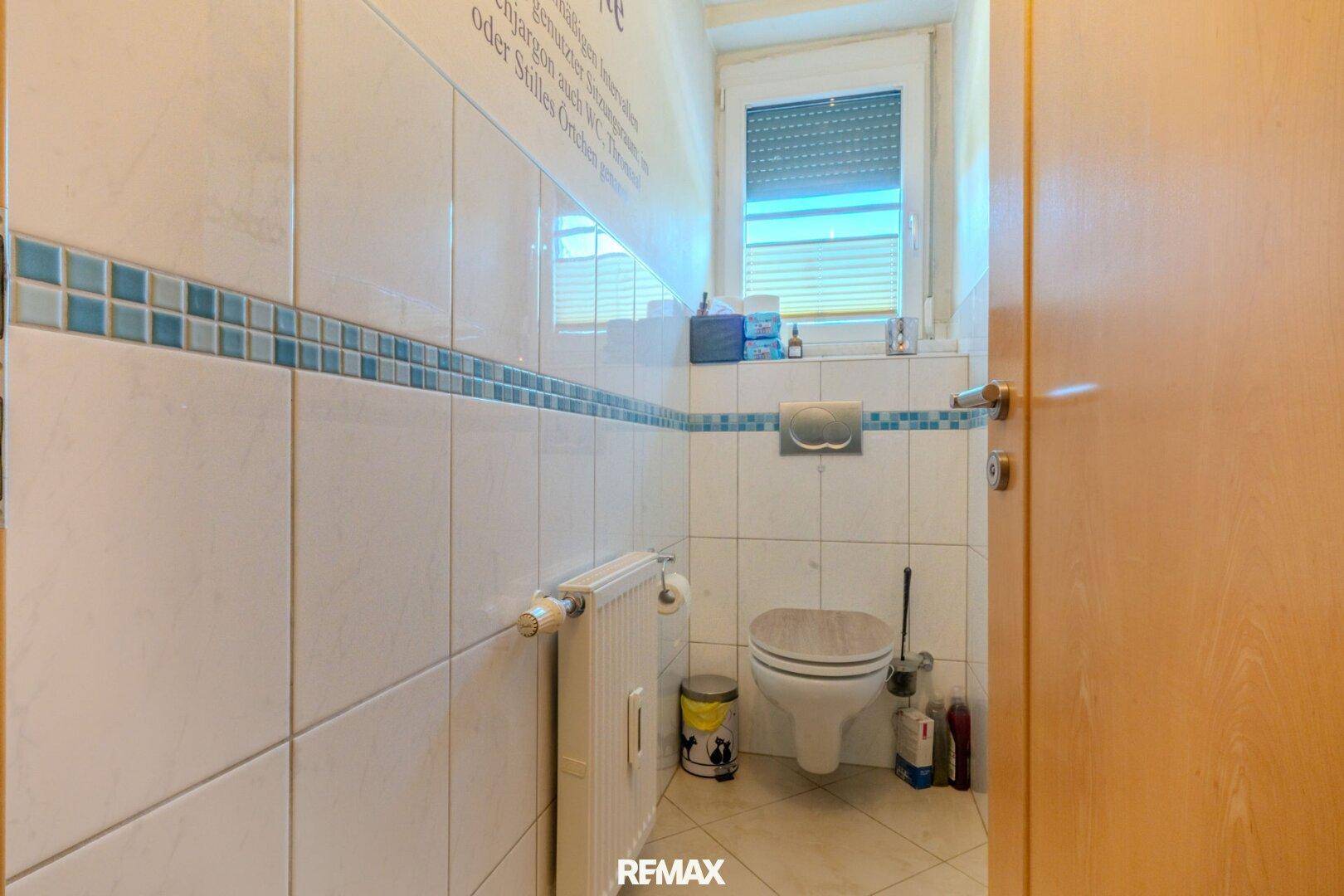Kleines WC mit weißen Fliesen, blauer Bordüre und Fenster.