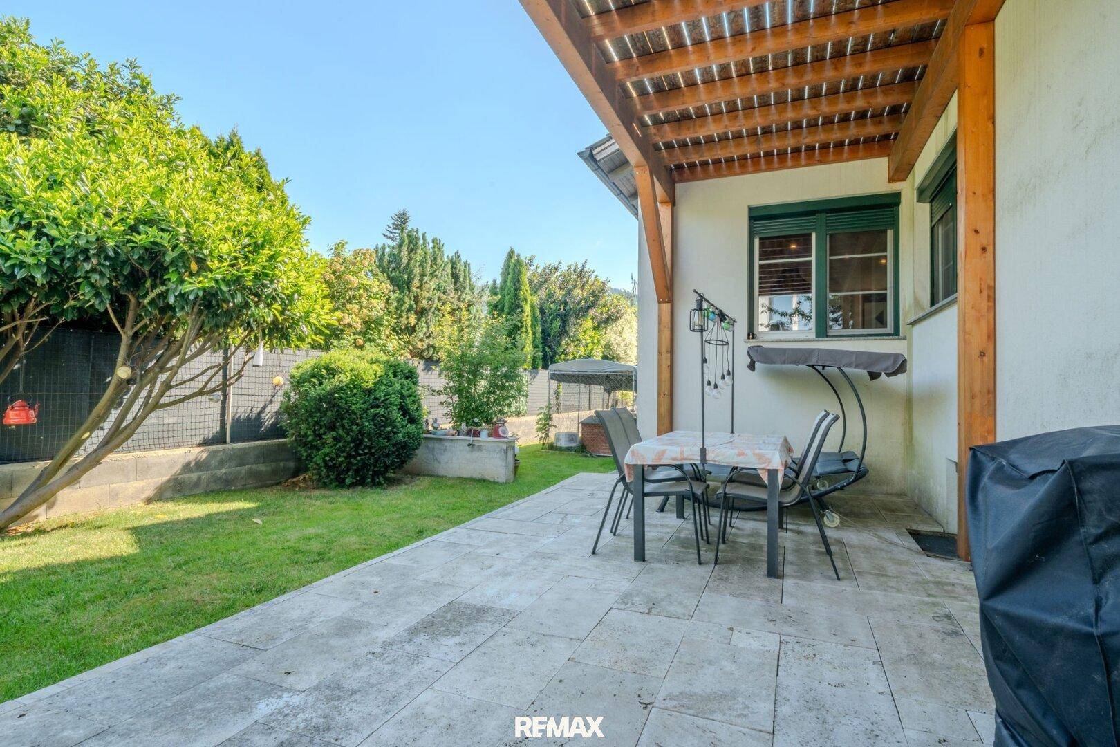 Gepflasterte Terrasse mit Gartenmöbeln, Pergola und angrenzendem Rasenbereich unter blauem Himmel.