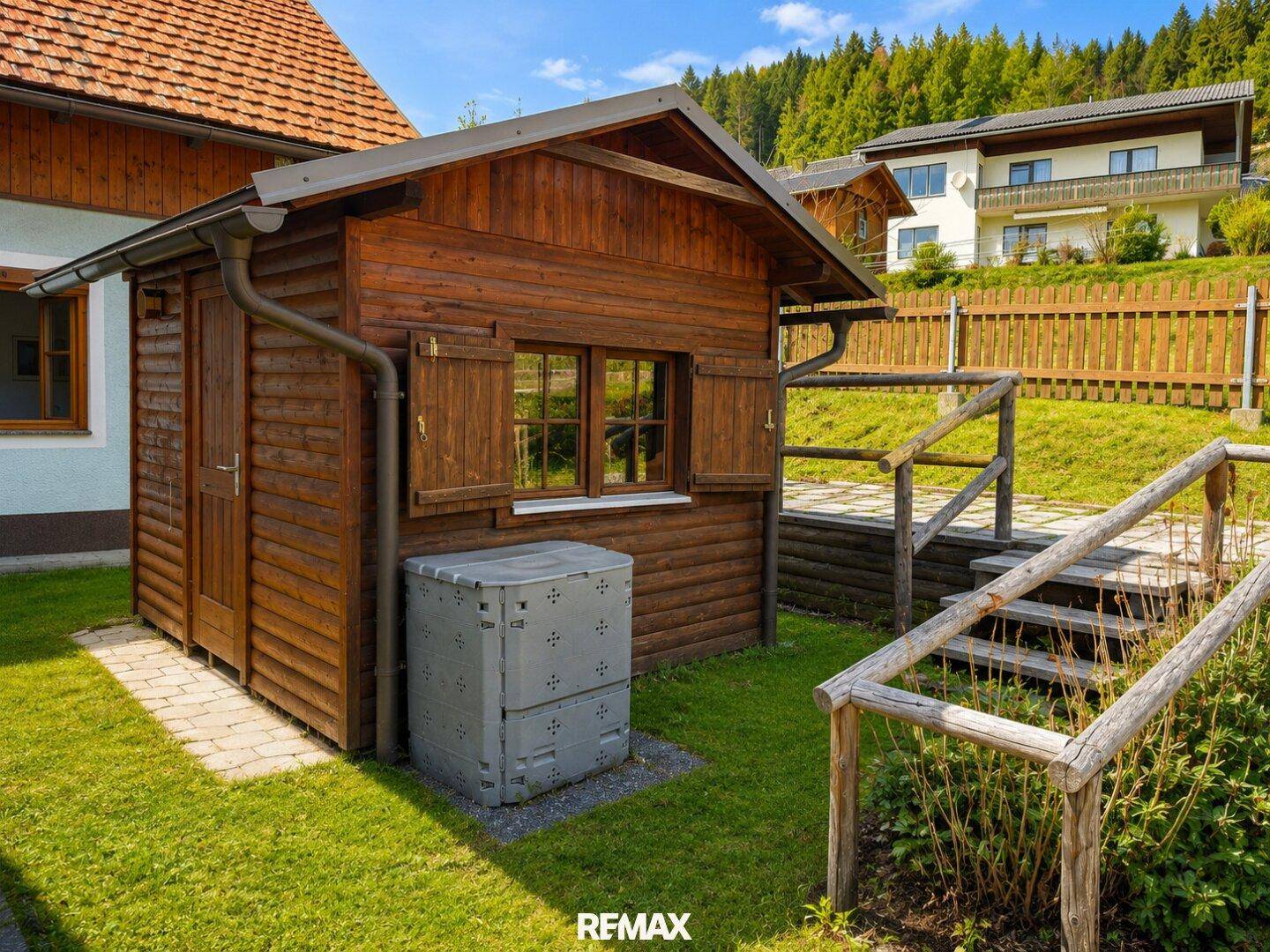 Kleines Holzgartenhaus mit Fensterläden und Komposter im grünen Gartenbereich.