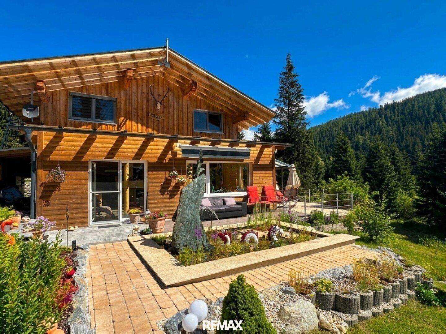 Holzhaus mit großer gepflasterter Terrasse, Wasserspiel und blühenden Blumenbeeten.