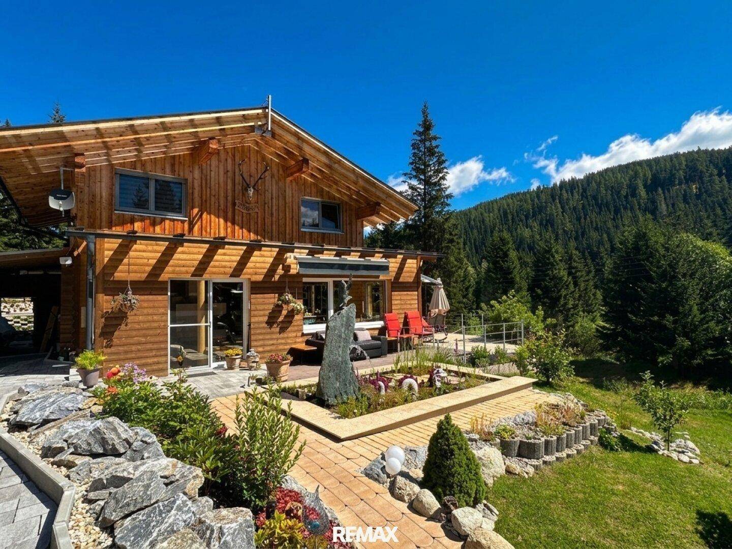 Geräumige Terrasse vor dem Holzhaus mit Wasserspiel, Gartenbeeten und Bergblick.
