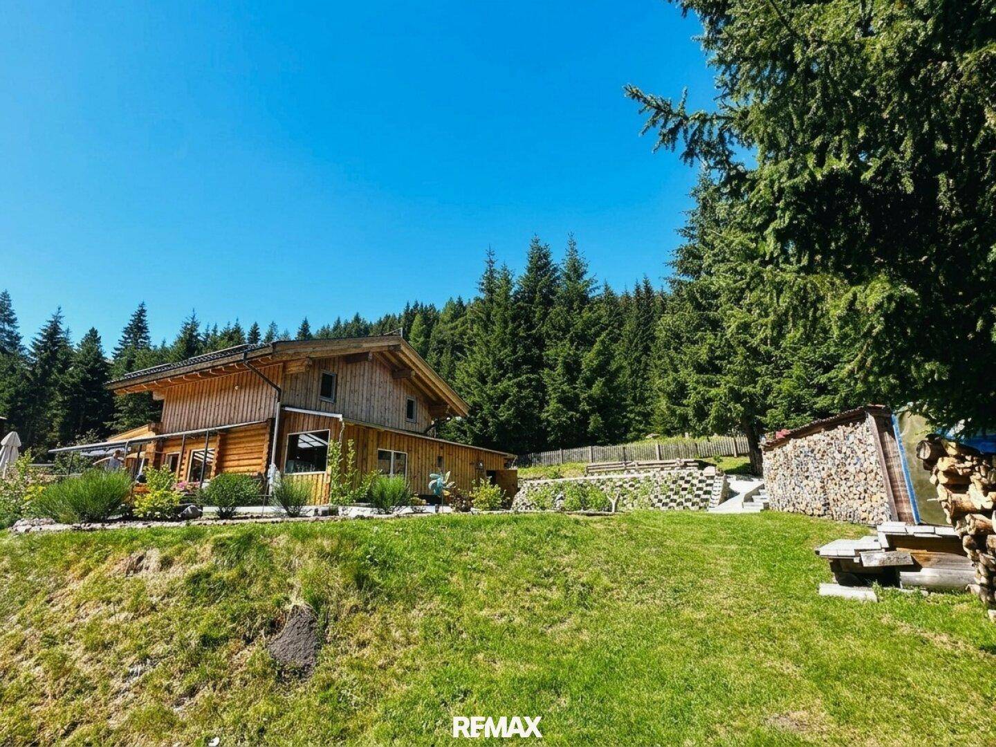 Seitenansicht des Holzhauses mit großem Holzstapel und einer grünen Hanglage.