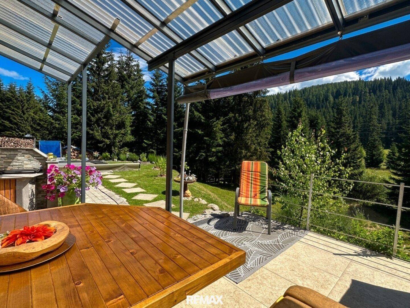Blick vom überdachten Essbereich auf die grüne Gartenlandschaft und den umliegenden Wald.