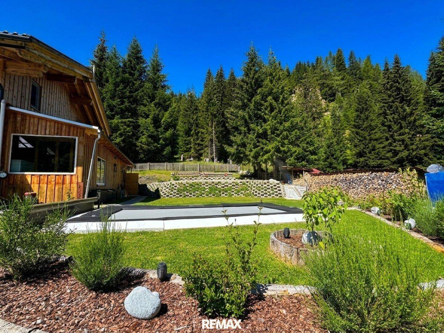 Gepflegter Gartenbereich mit überdachtem Trampolin, Sträuchern und dem Holzhaus im Hintergrund.