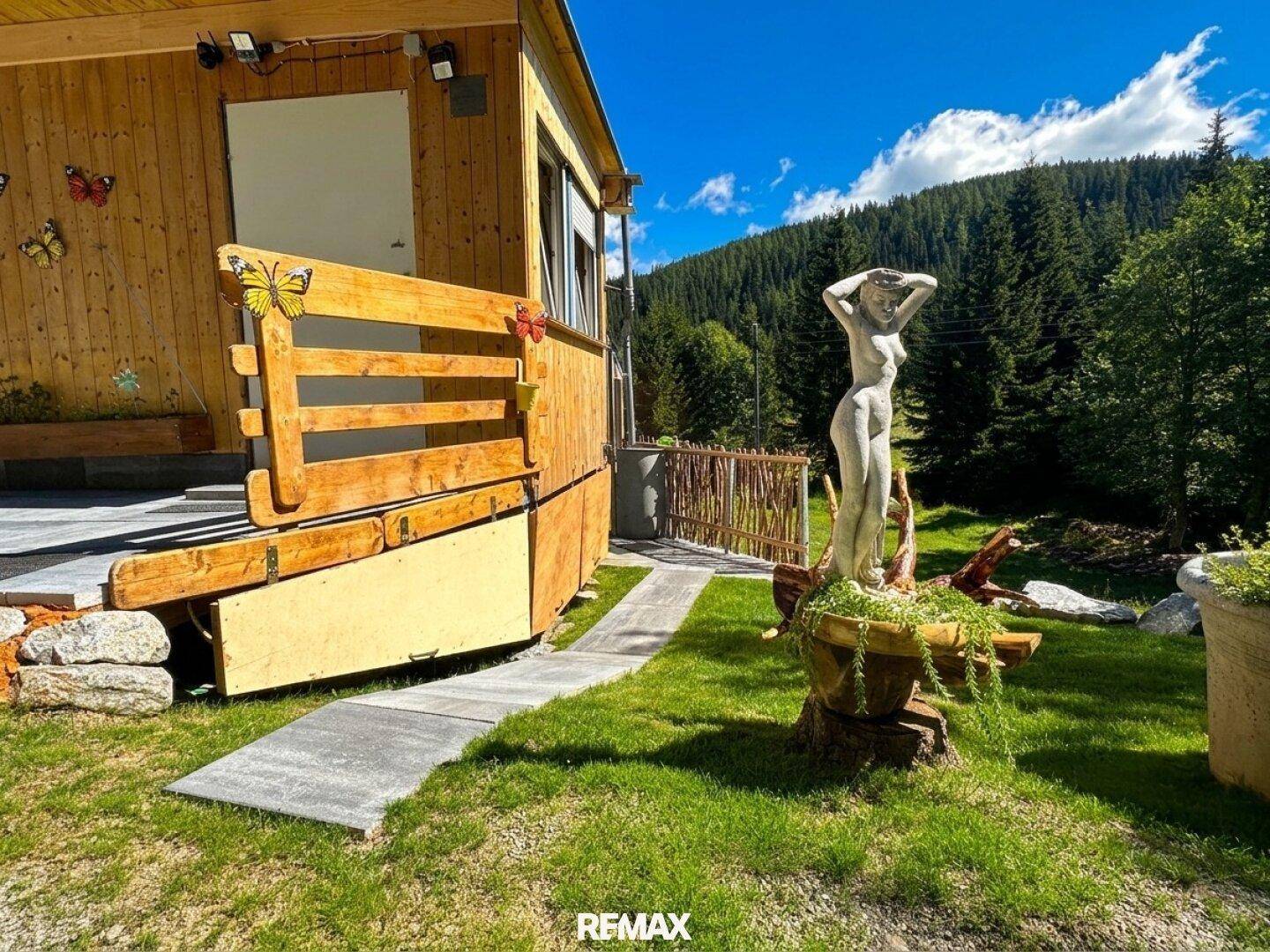 Holzhausfassade mit gepflastertem Weg, einer Statue und Blick auf den umliegenden Wald.