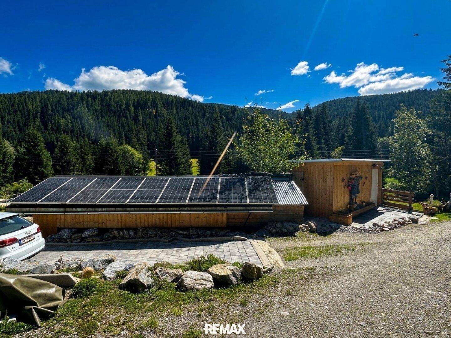 Holzgebäude mit Solaranlage und angrenzendem Schuppen in bergiger Landschaft unter blauem Himmel.