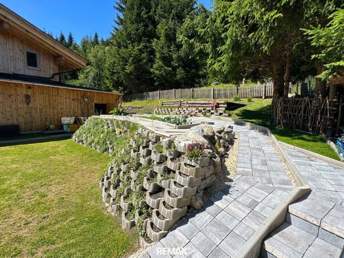 Gepflasterter Weg mit Stufen und bepflanzter Stützmauer, führt zu terrassierten Gartenbeeten.