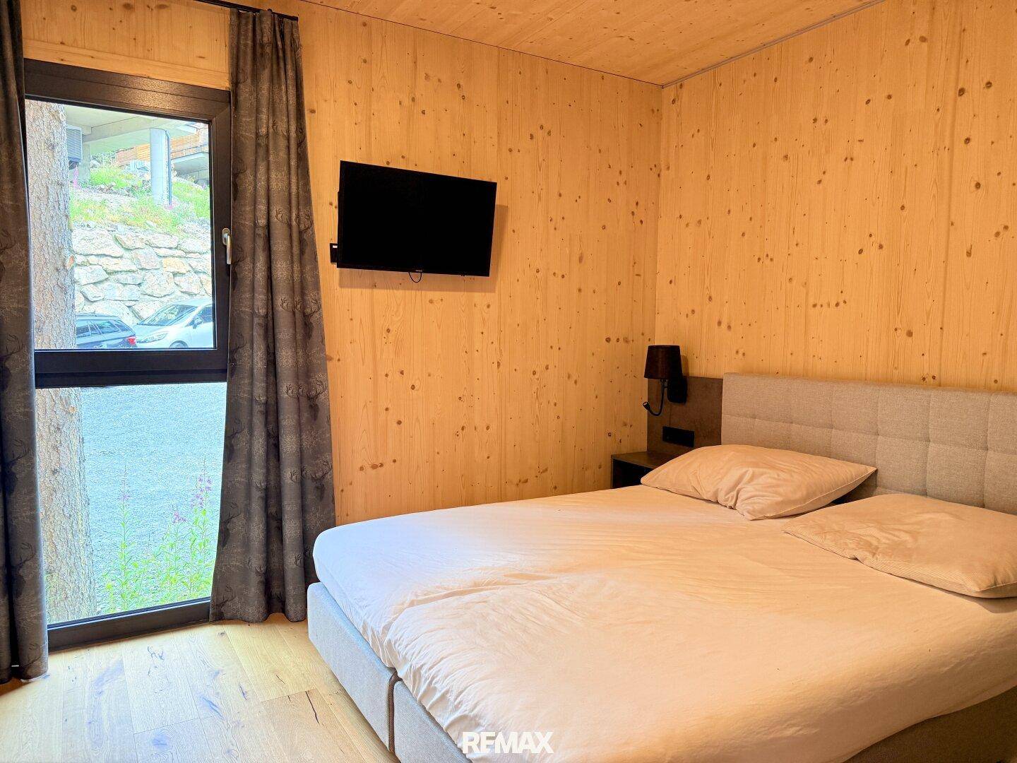 Gemütliches Schlafzimmer mit Holzwänden, Doppelbett und großem Fenster mit Blick ins Grüne.