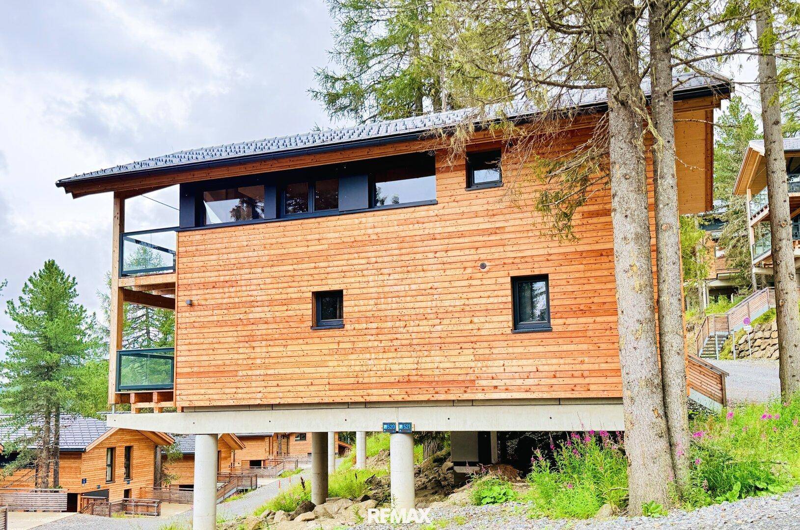 Seitliche Ansicht eines modernen Holzhauses auf Stelzen mit großen Fenstern und Balkon.