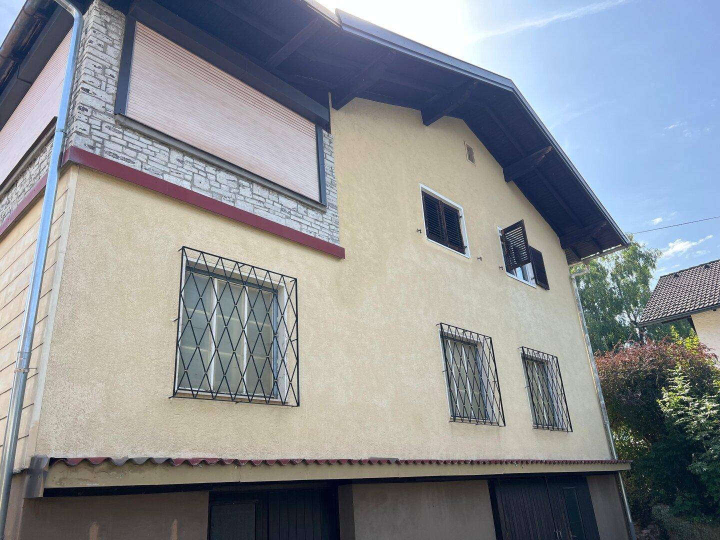 Seitenansicht des Hauses mit gelber Fassade, Fenstergittern und Rollläden unter hellem Himmel.