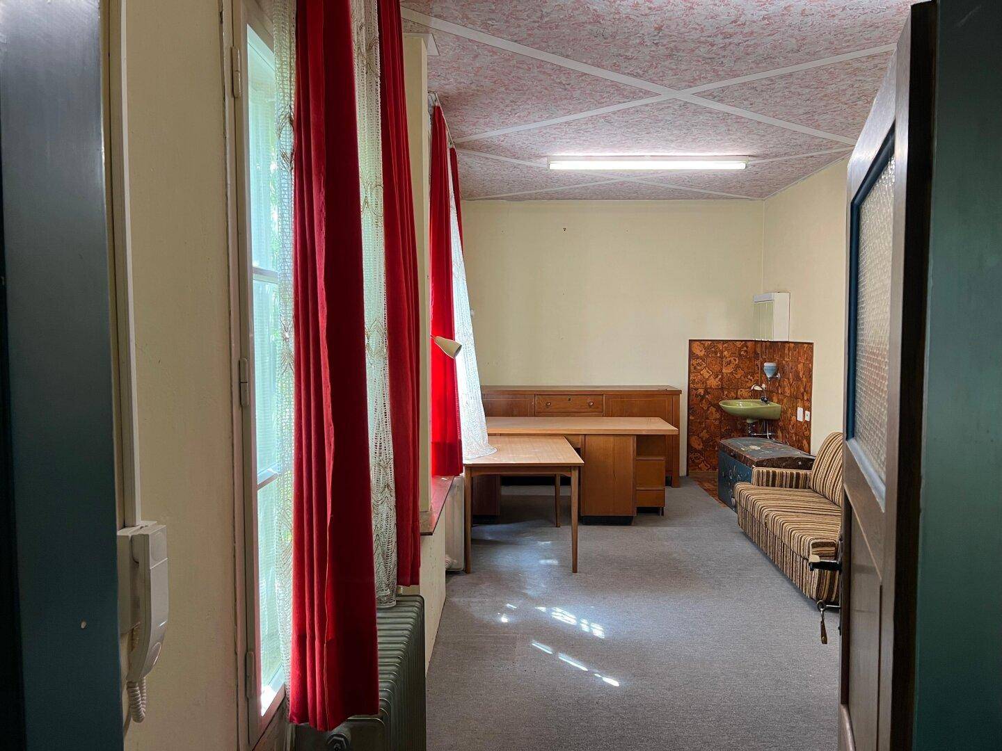 Innenraum mit Schreibtisch, Sofa und großen Fenstern mit roten Vorhängen, im älteren Stil.
