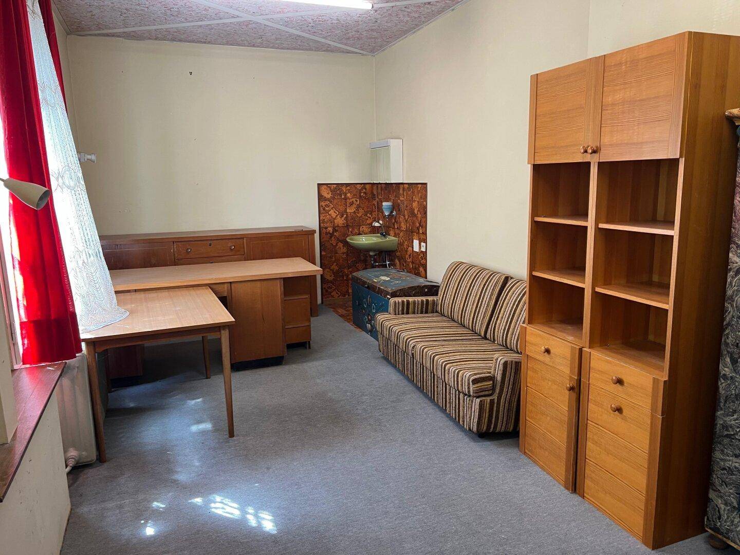 Innenraum mit Holzschränken, Sofa und Schreibtisch, geprägt von älterer Einrichtung und Teppichboden.