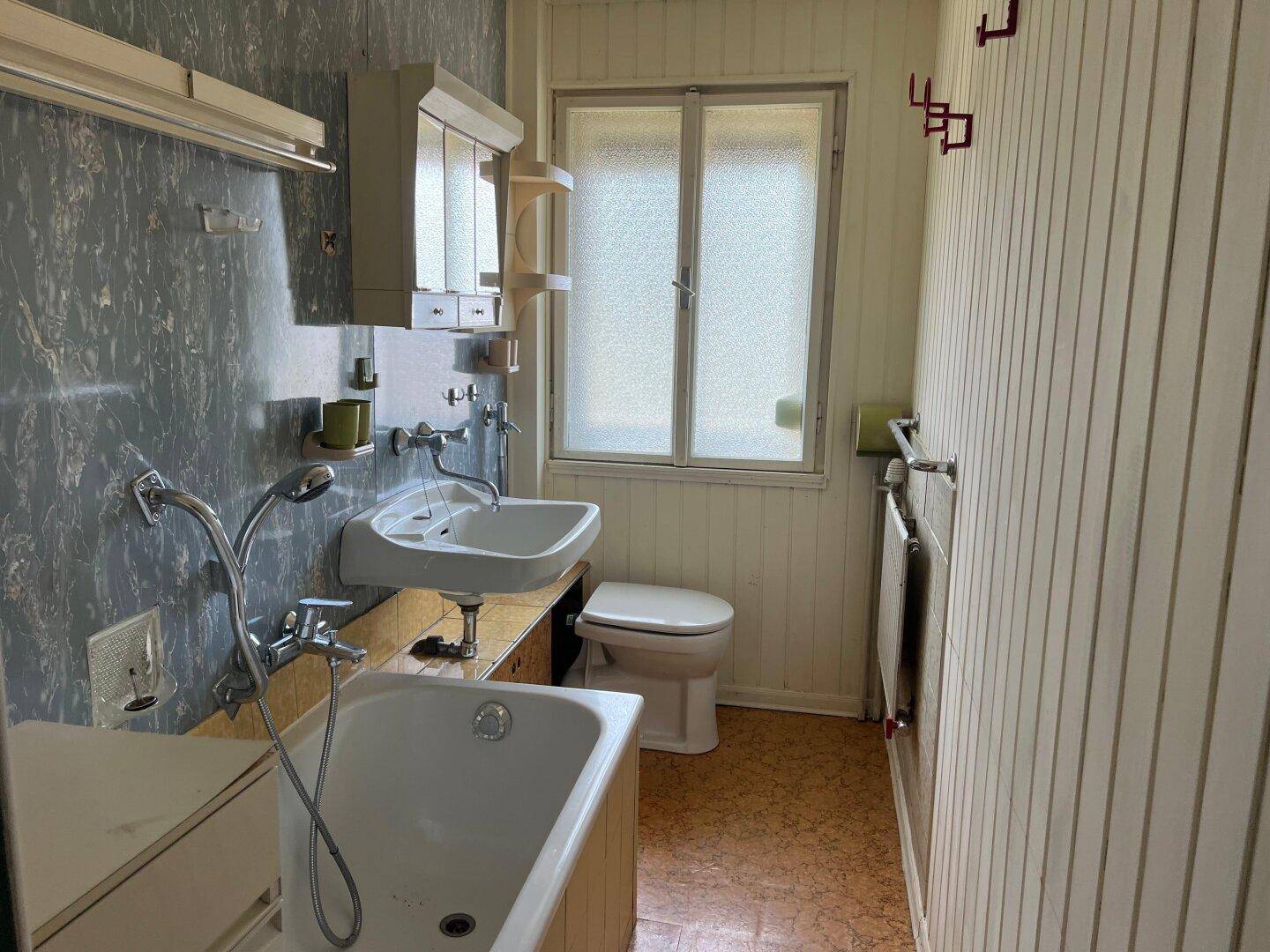 Badezimmer mit Badewanne, Waschbecken und Fenster, im älteren Stil gehalten und renovierungsbedürftig.