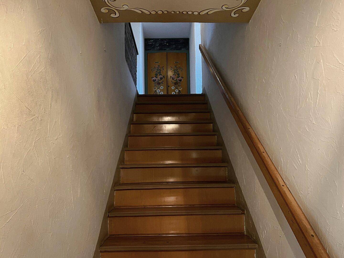 Holztreppe führt zu einem oberen Stockwerk, mit einfacher Wandgestaltung und Holzgeländer.
