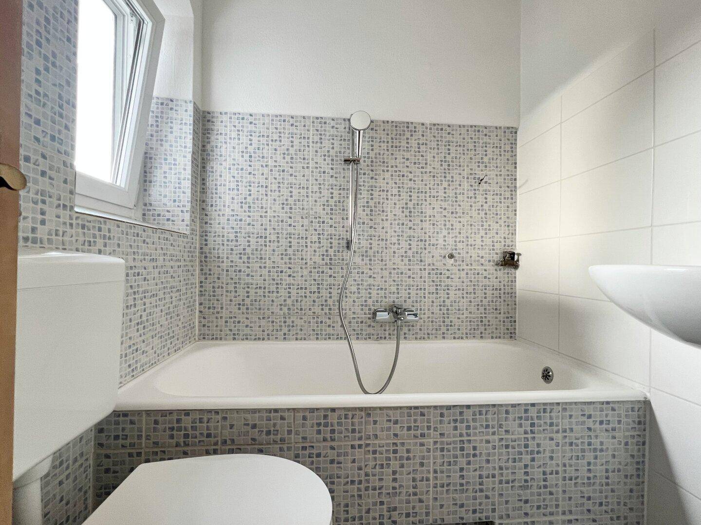 Badezimmer mit Badewanne, WC und Mosaikfliesen an den Wänden, hell durch ein Fenster.