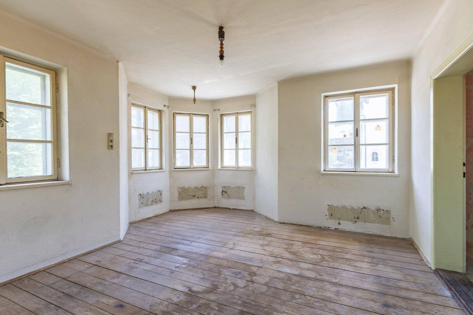 Leerer Raum mit Holzdielenboden und vielen Fenstern, der auf eine Renovierung wartet.