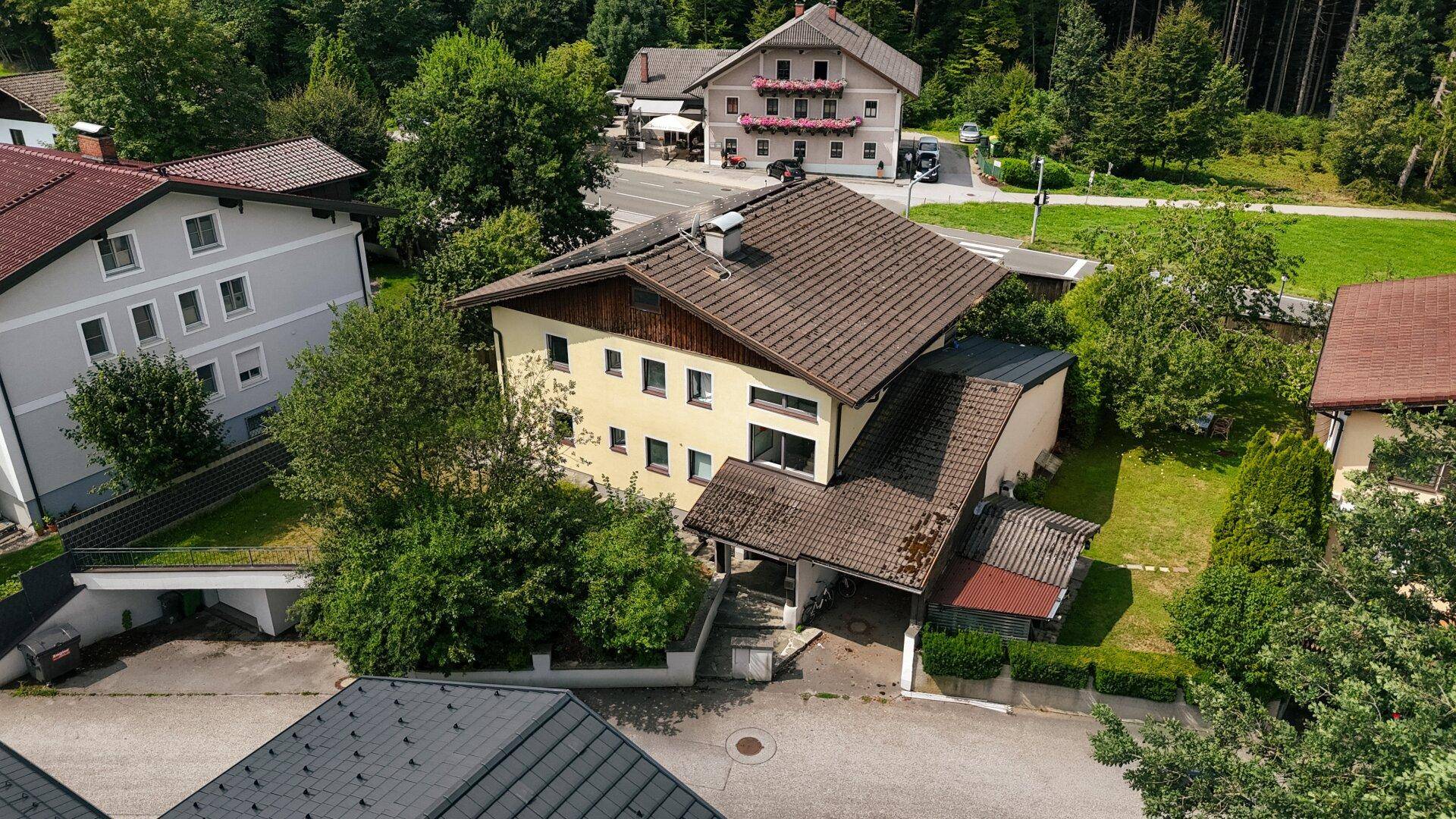Schrägansicht des gelben Hauses mit braunem Dach, eingebettet in eine grüne Umgebung mit weiteren Gebäuden.