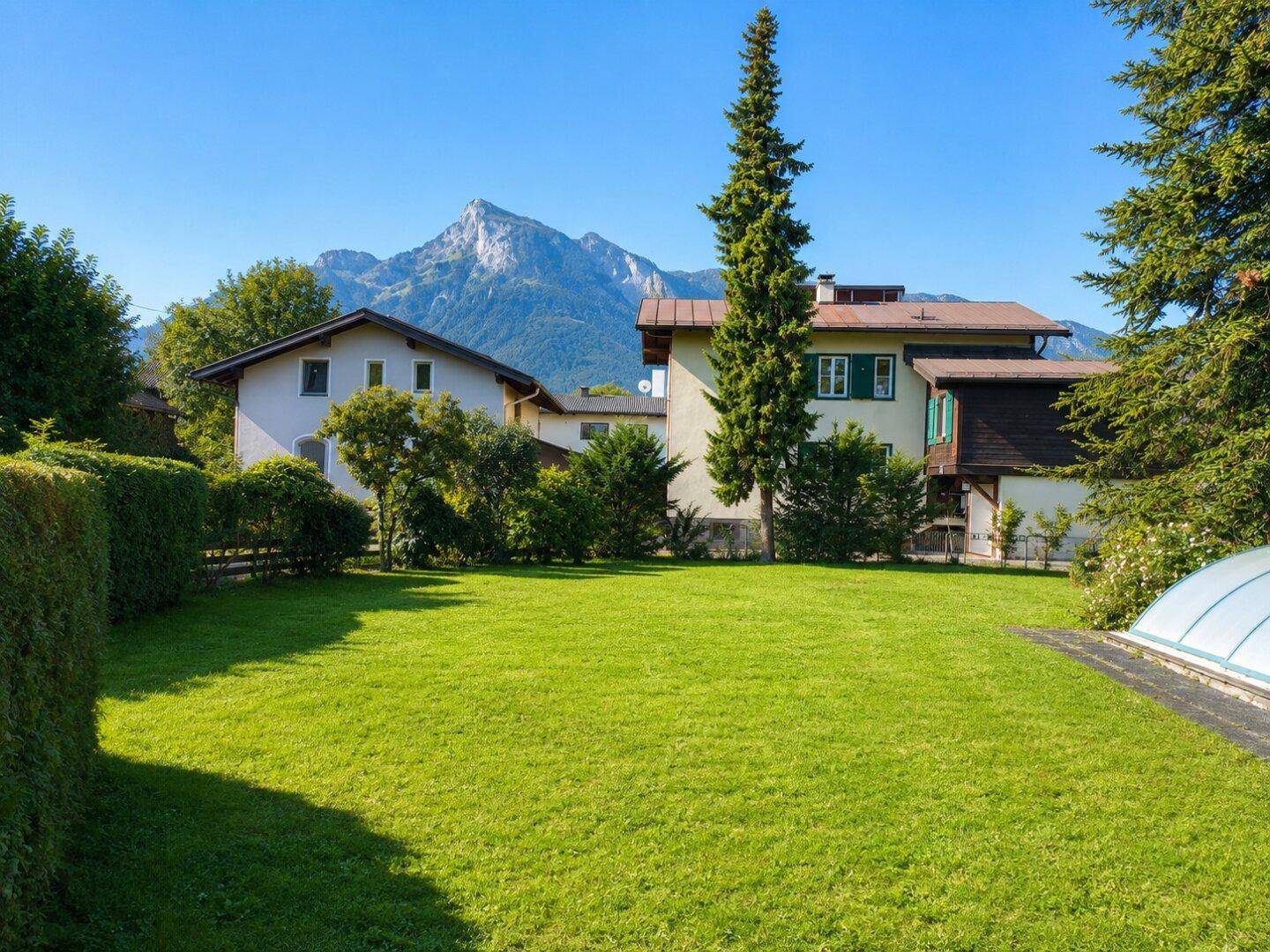 Sonniger, weitläufiger Garten mit Rasenfläche, umgeben von Hecken und Bäumen, mit Bergblick.