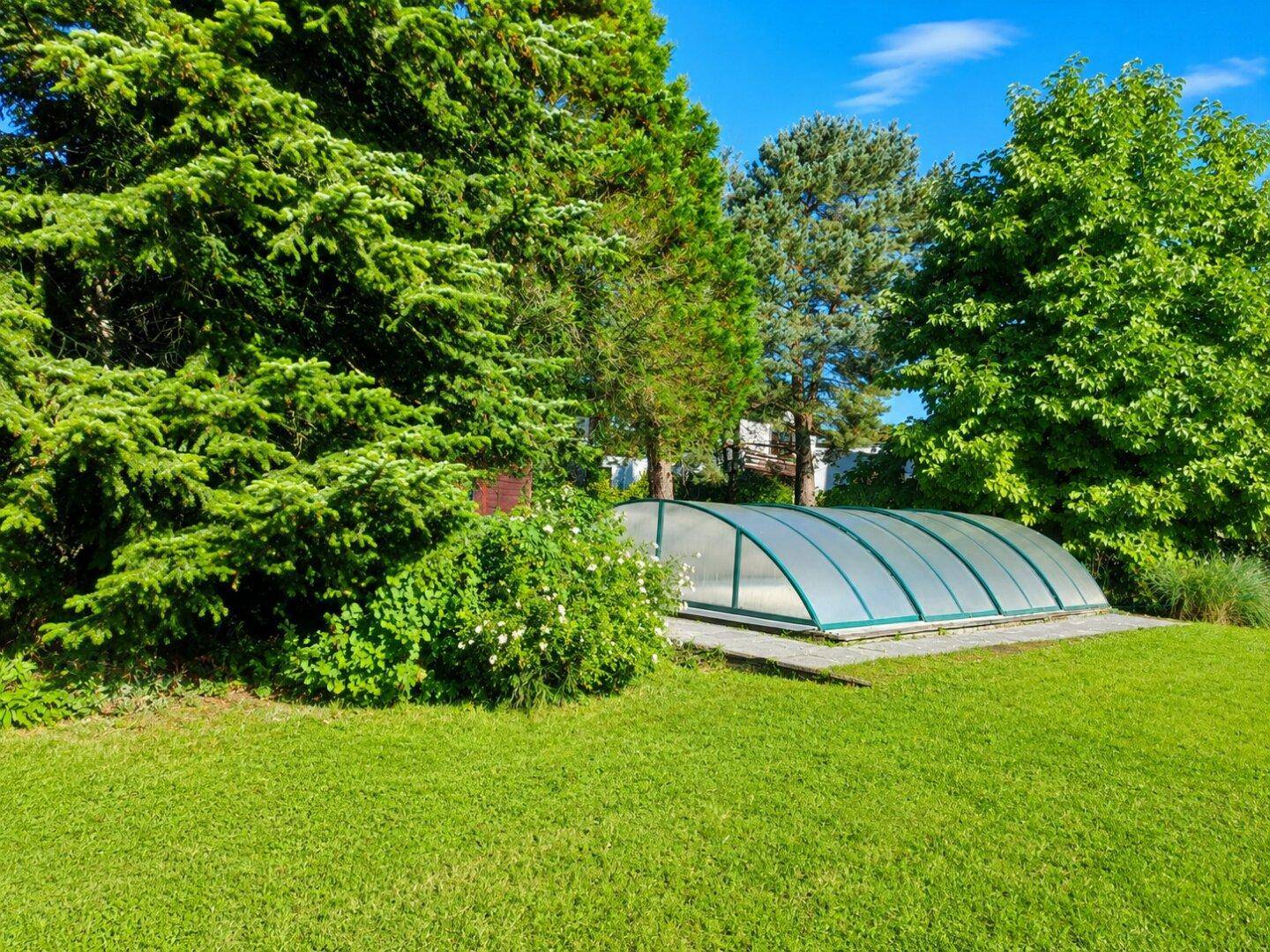 Überdachter Poolbereich im grünen Garten, umgeben von hohen Bäumen und üppiger Vegetation.