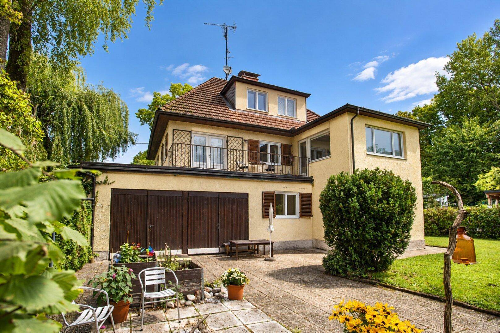 Gelbes Einfamilienhaus mit Satteldach, Balkon und integrierter Garage, umgeben von einem grünen Garten.