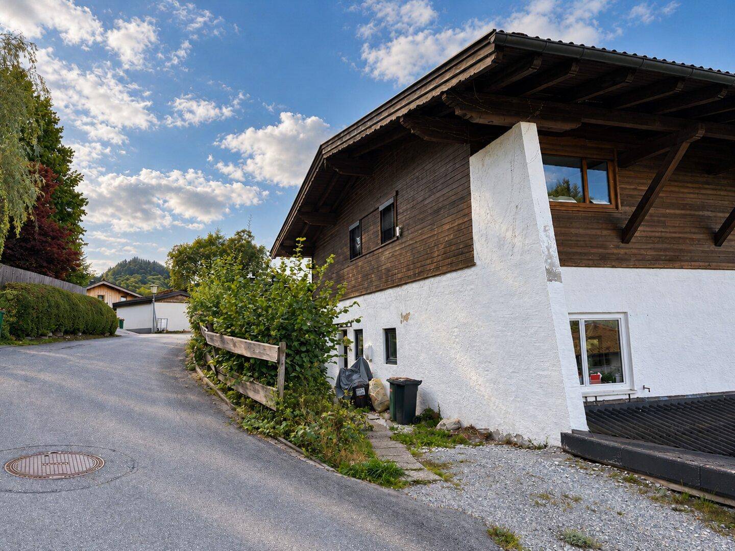 Traditionelles Haus mit Holzfassade und weißem Sockel an einer leicht ansteigenden Straße.