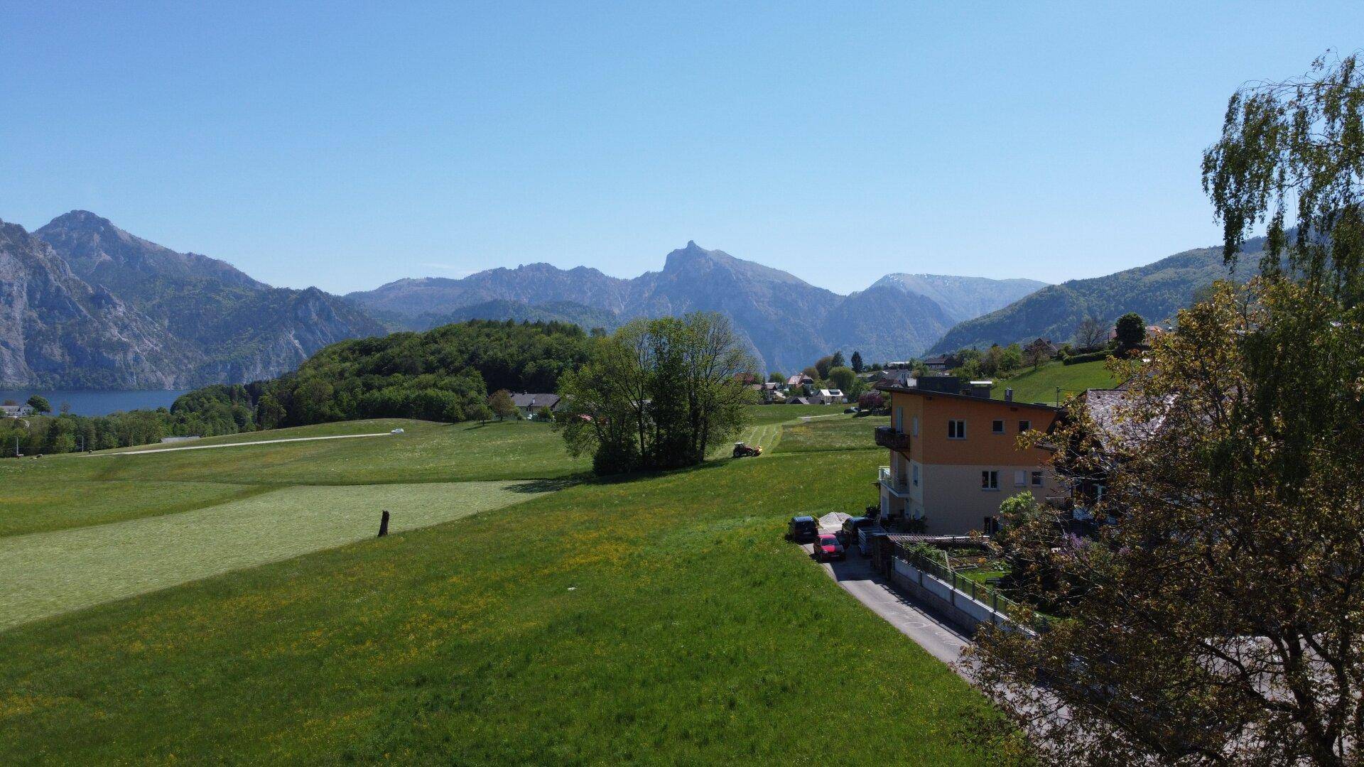 Blick über grüne Hügel und Häuser auf eine beeindruckende Bergkette und einen See in der Ferne.