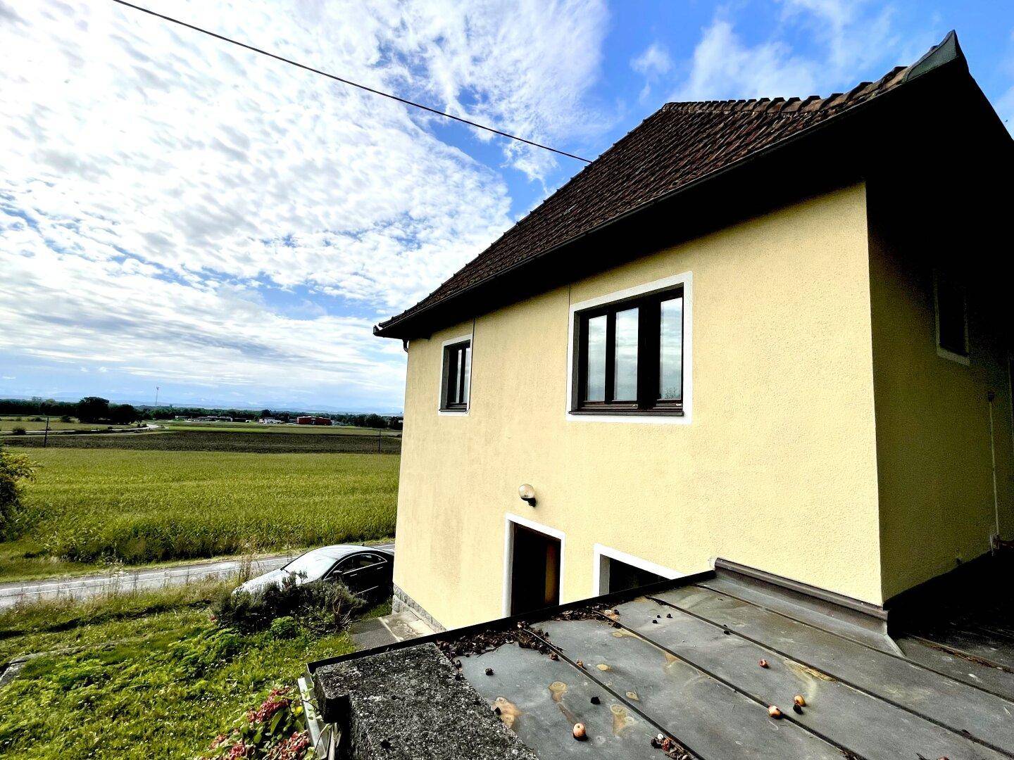 Seitenansicht des Hauses mit gelber Fassade und Blick über ein Feld unter einem bewölkten Himmel.