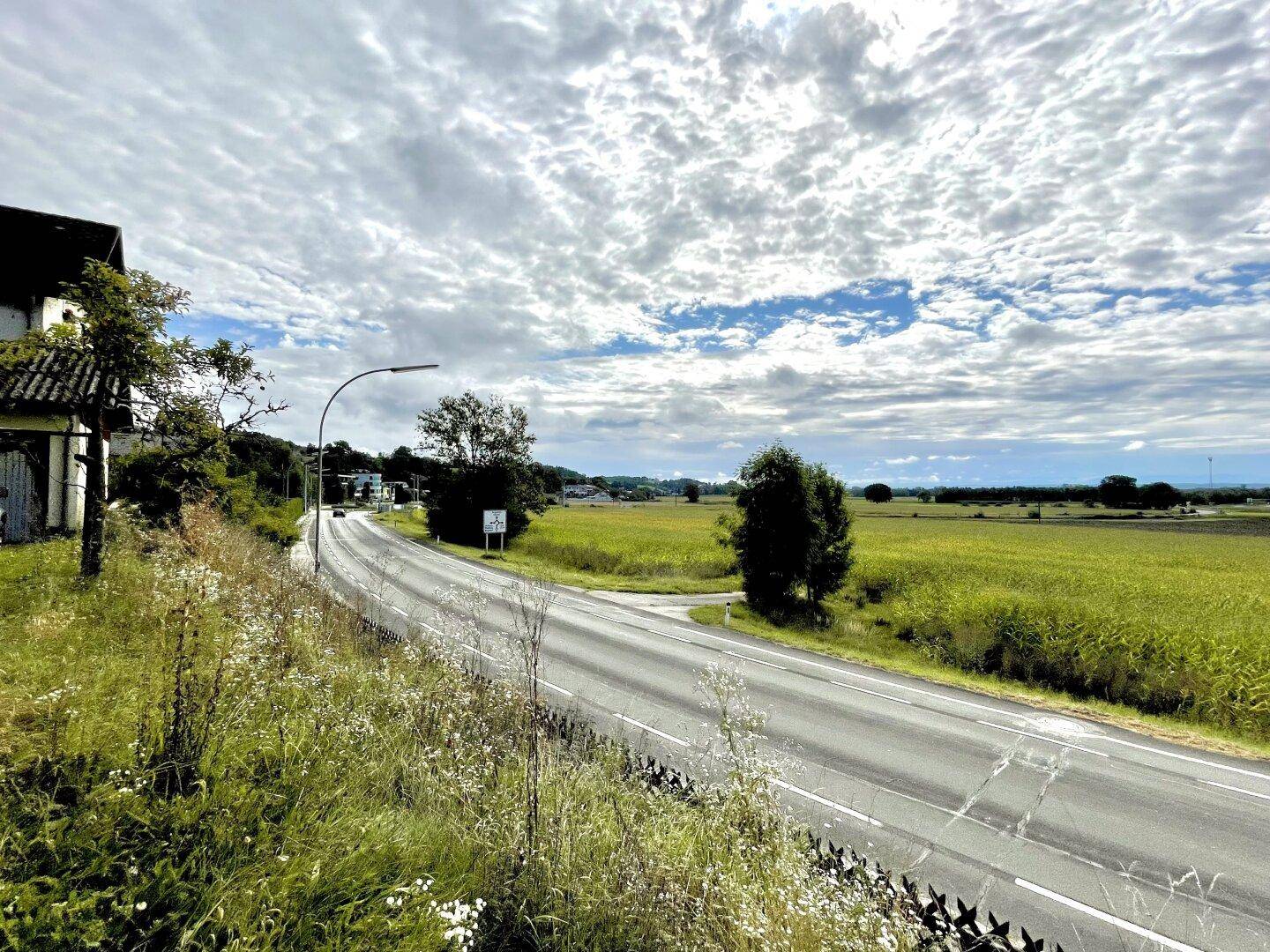 Weitläufige Landschaft mit einer Landstraße, umgeben von grünen Feldern unter einem dramatischen Himmel.