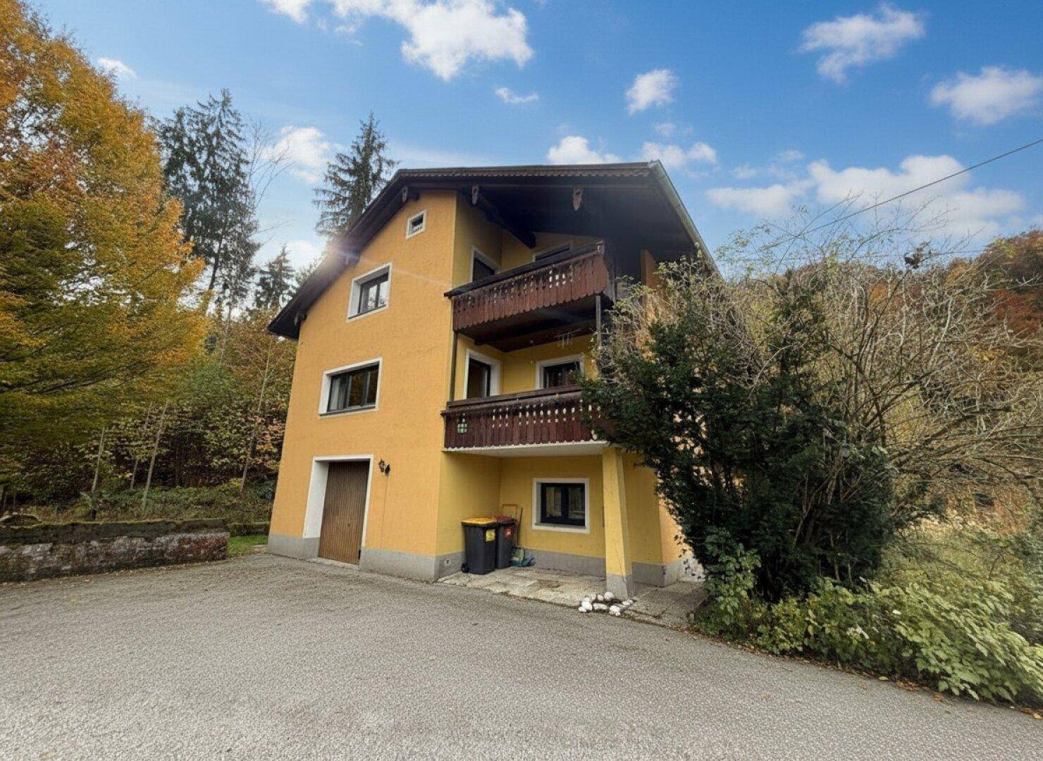 Vorderansicht eines gelb verputzten Hauses mit Holzbalkonen und umgeben von Herbstbäumen.