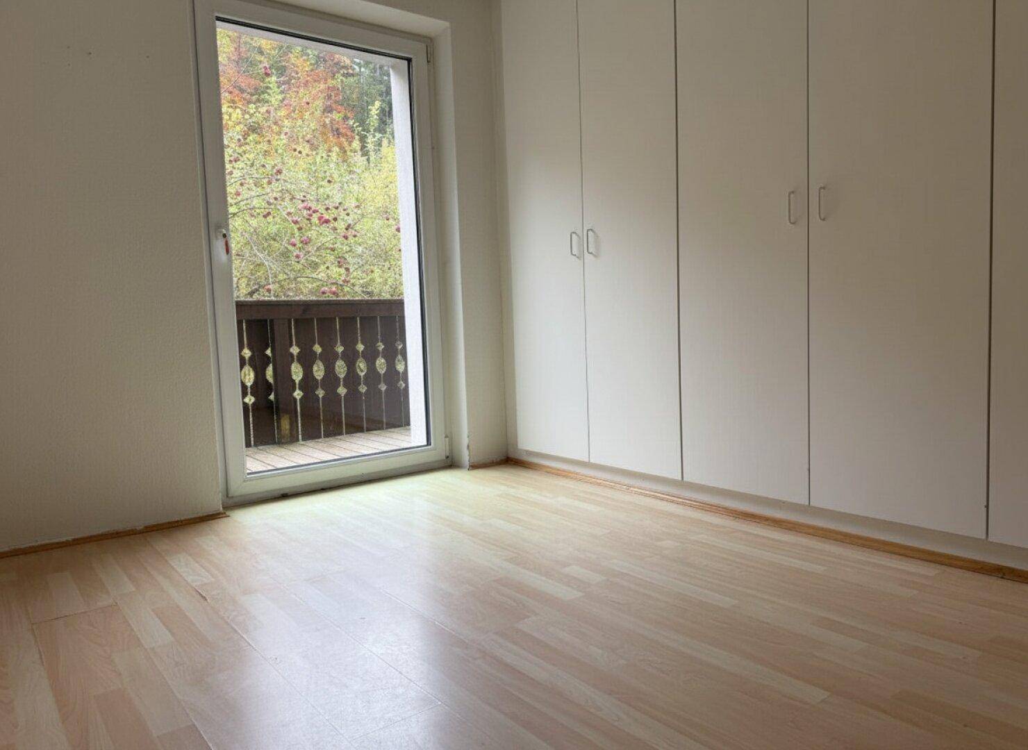 Schlafzimmer mit hellen Einbauschränken, Laminatboden und Balkonzugang mit Waldblick.