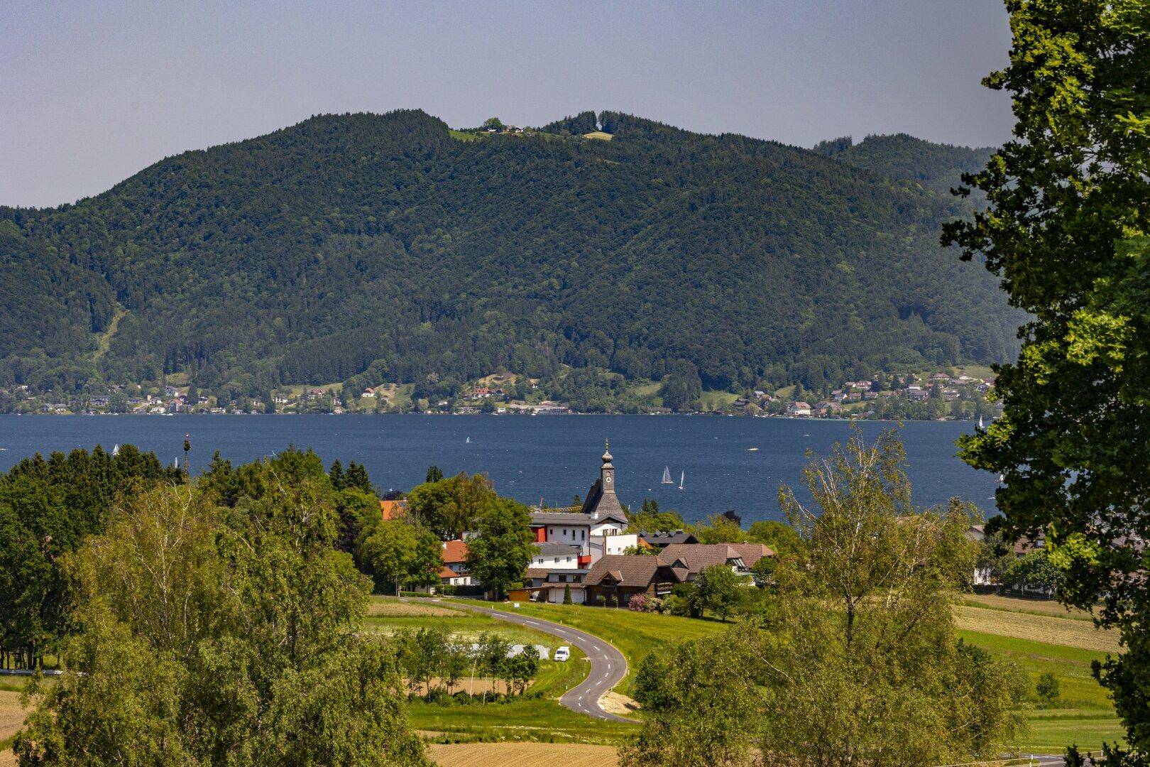 Weitläufiger Blick auf ein idyllisches Dorf am See mit grünen Hügeln und Bergen.
