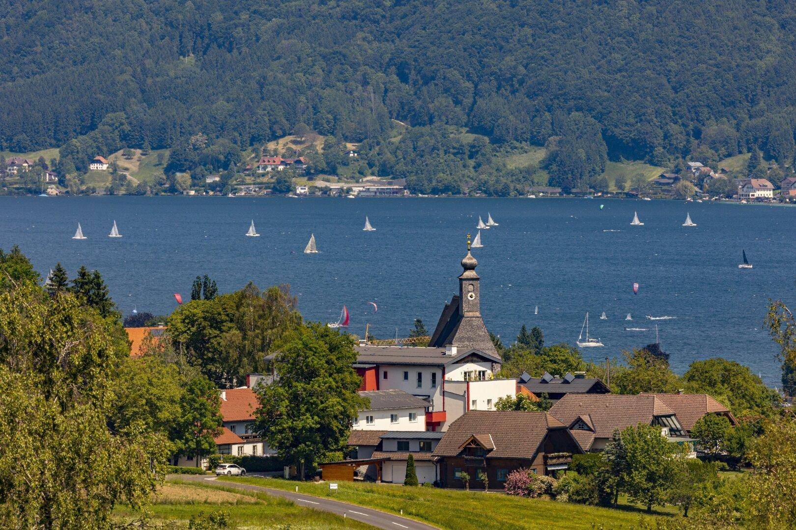 Malerisches Dorf am Seeufer mit Segelbooten und bewaldeten Bergen im Hintergrund.
