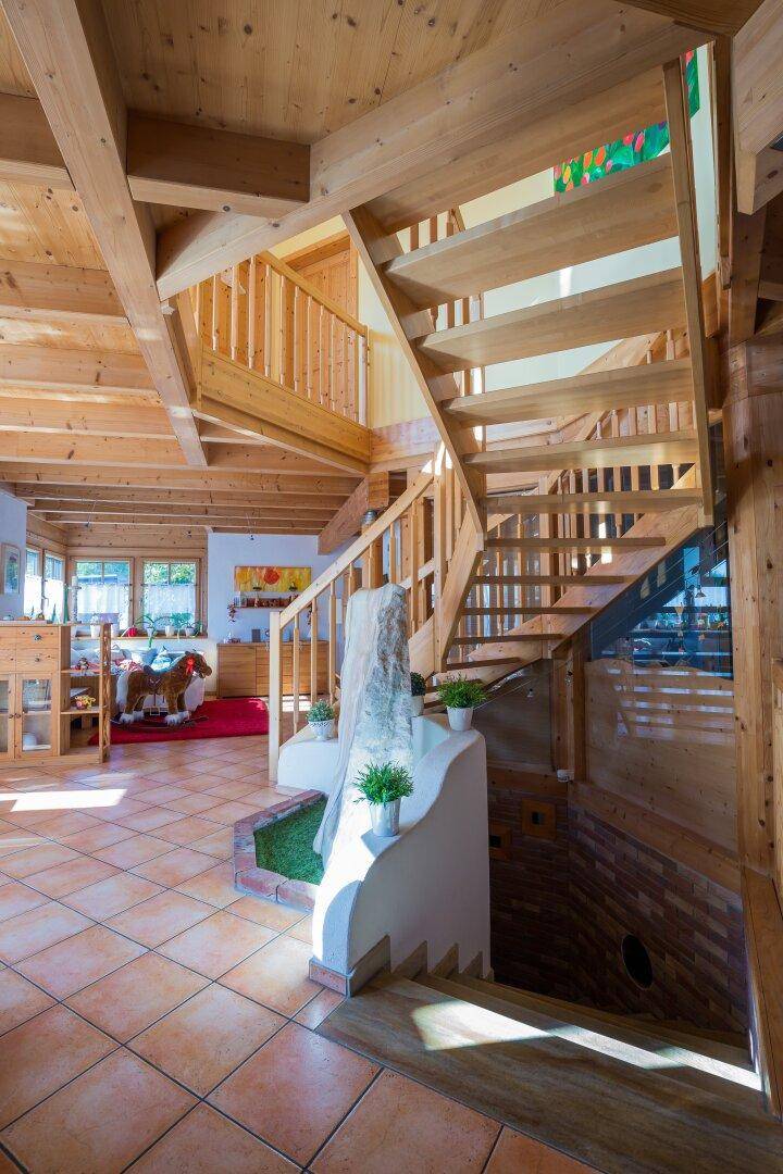 Heller Eingangsbereich mit Holztreppe, Terrakottafliesen und sichtbaren Holzbalken, der zu den oberen Etagen führt.