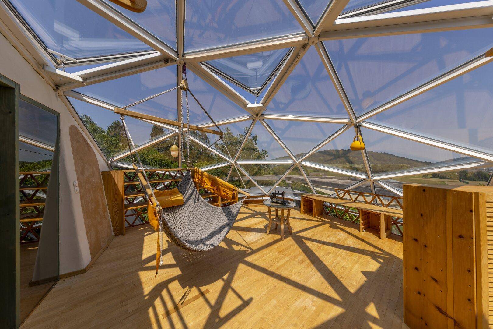 Heller Raum unter einer Glaskuppel mit Holzboden und Hängematte, bietet weite Ausblicke.