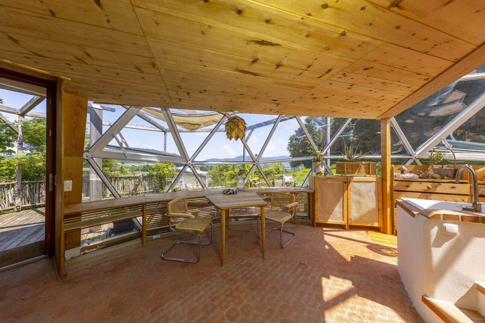 Gemütlicher Essbereich mit Holztisch und Stühlen unter einer Glaskonstruktion mit Panoramablick.