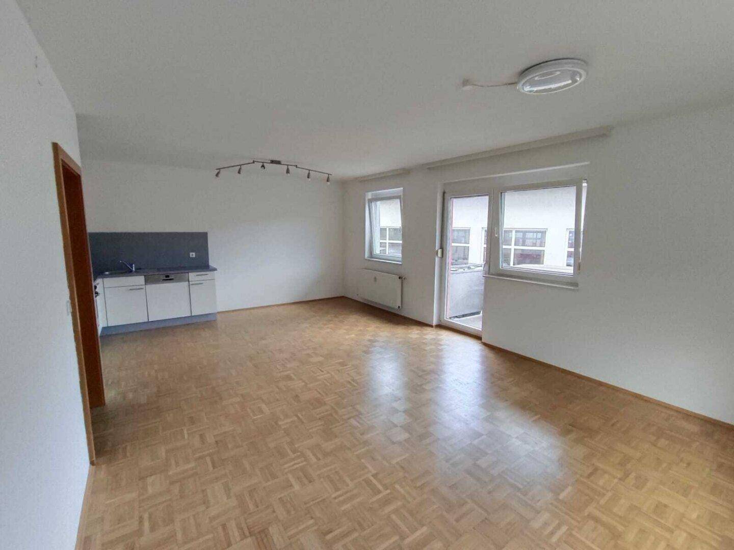 Heller Wohnbereich mit Parkettboden, kleiner Küchenzeile und Zugang zum Balkon.