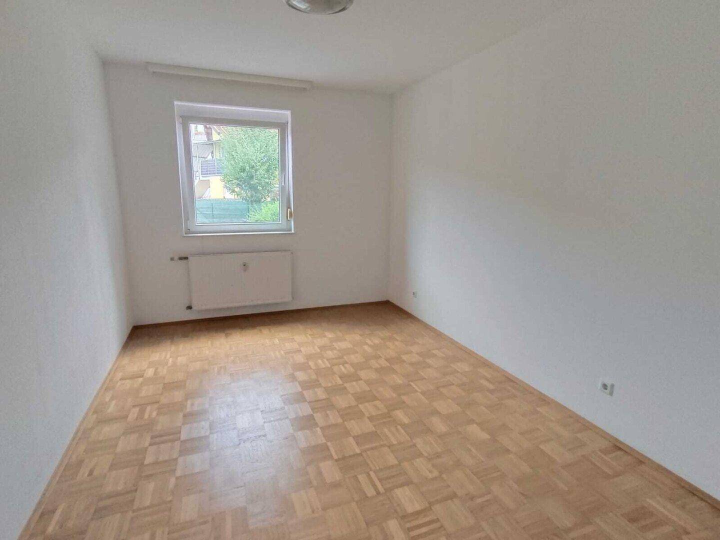 Helles Schlafzimmer mit Parkettboden und einem Fenster mit Blick ins Grüne.