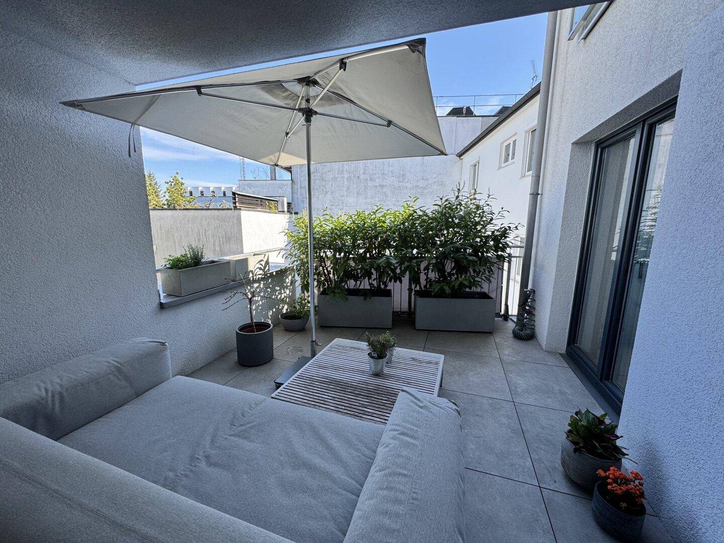 Sonniger Außenbereich mit Lounge-Möbeln, Sonnenschirm und Pflanzen in Kübeln.