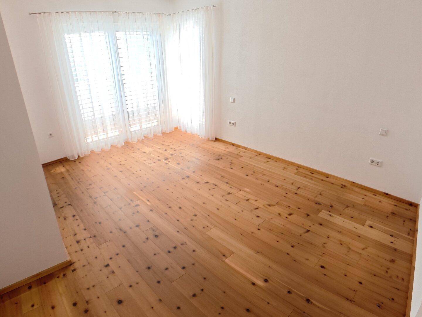 Leerer Raum mit hellem Holzboden und großen Fenstern, die viel Tageslicht hereinlassen.