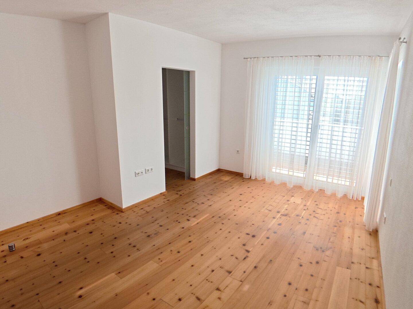 Unmöblierter Raum mit Holzdielenboden und breiter Fensterfront, die den Raum hell erscheinen lässt.