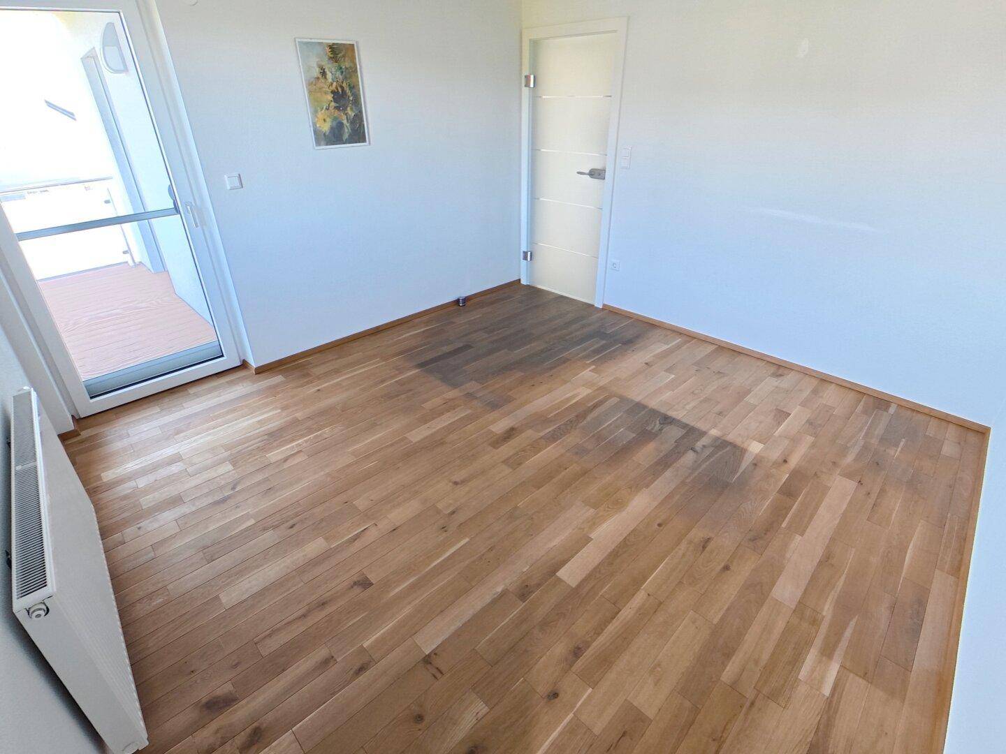 Leeres Zimmer mit hellem Holzparkettboden, Zugang zu einem Balkon und viel Tageslicht.
