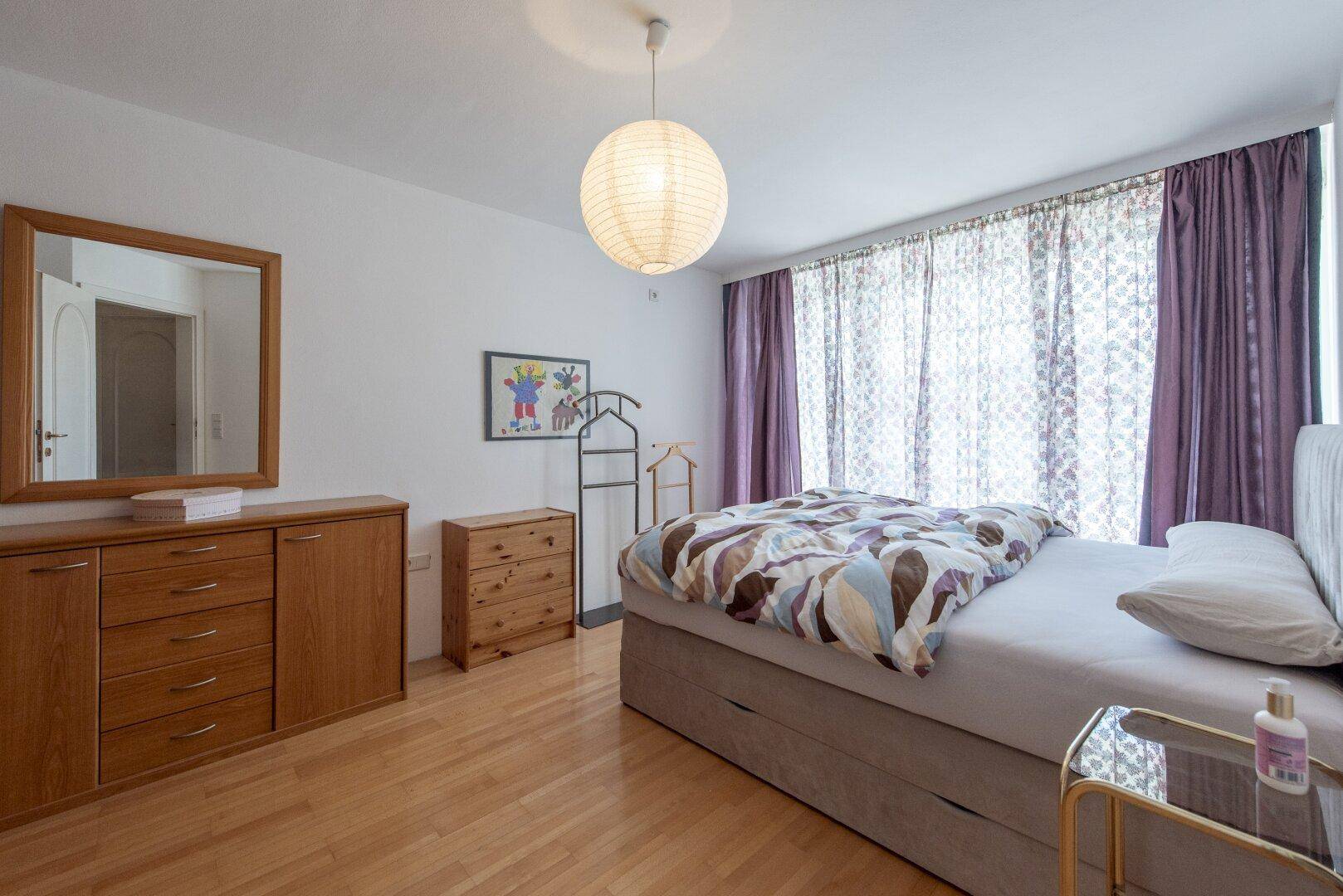 Schlafzimmer mit Holzboden, großem Bett und Vorhängen, die viel Licht hereinlassen.