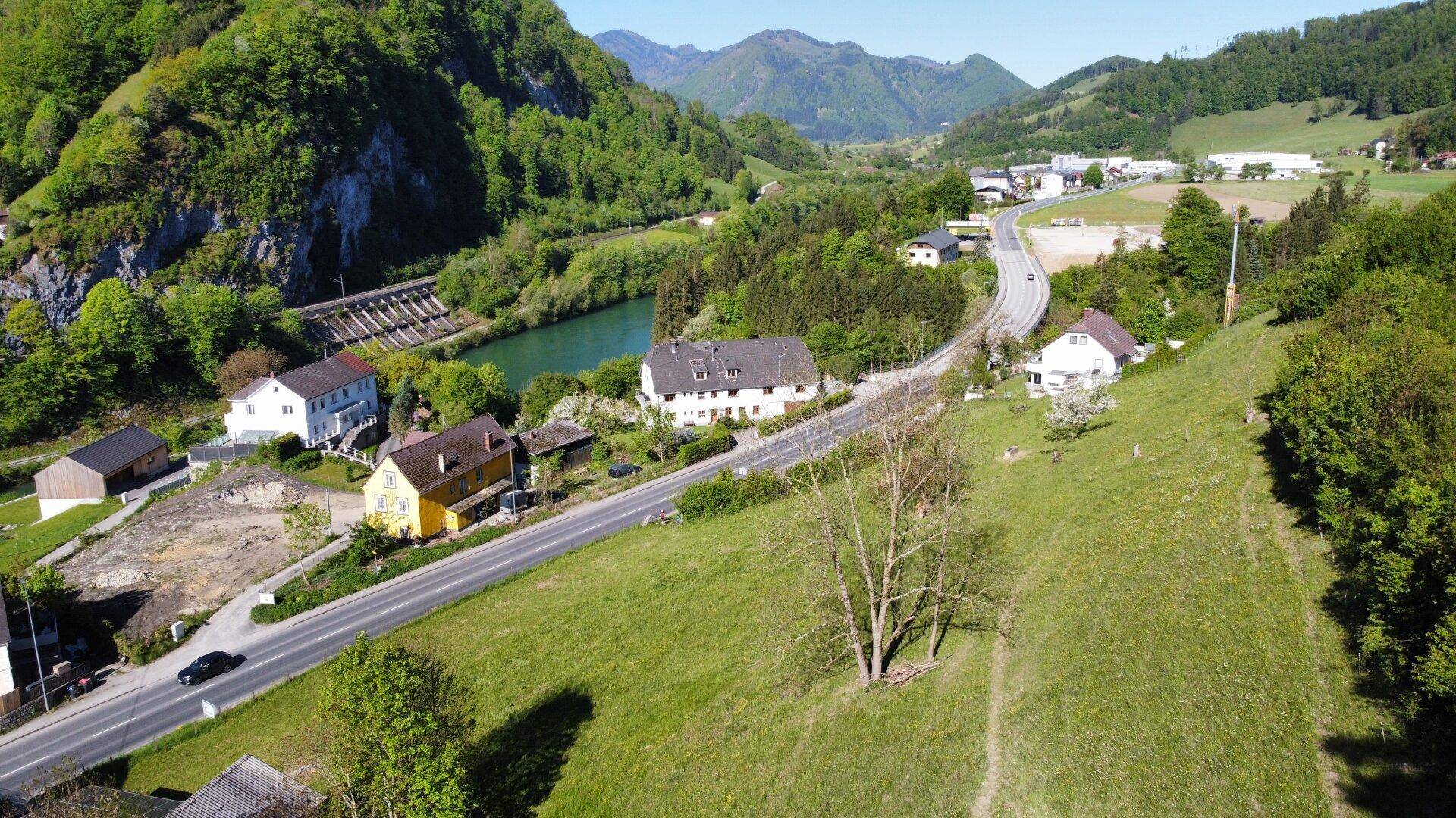 Weitläufiger Blick über einen grünen Hang mit einem Fluss, Häusern und einer Staumauer.