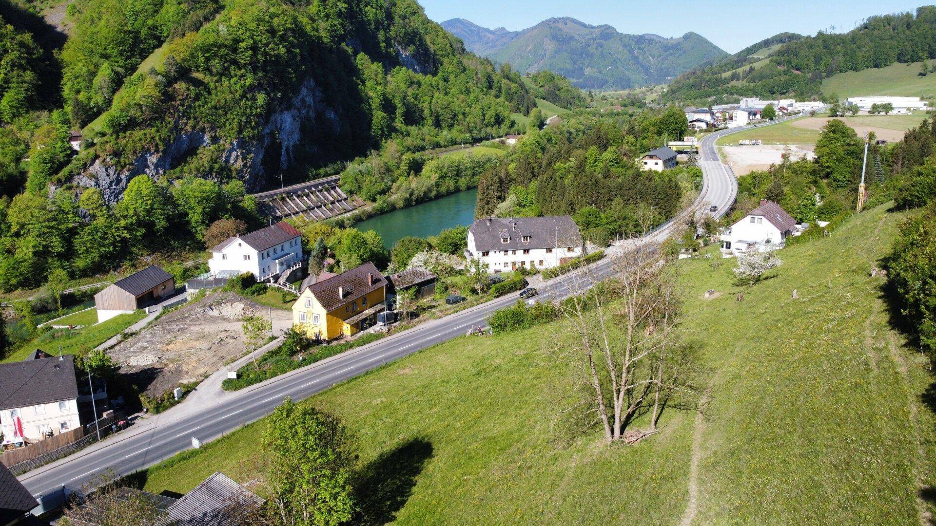 Panoramablick auf eine grüne Landschaft mit einem Fluss, Häusern und bewaldeten Bergen im Hintergrund.