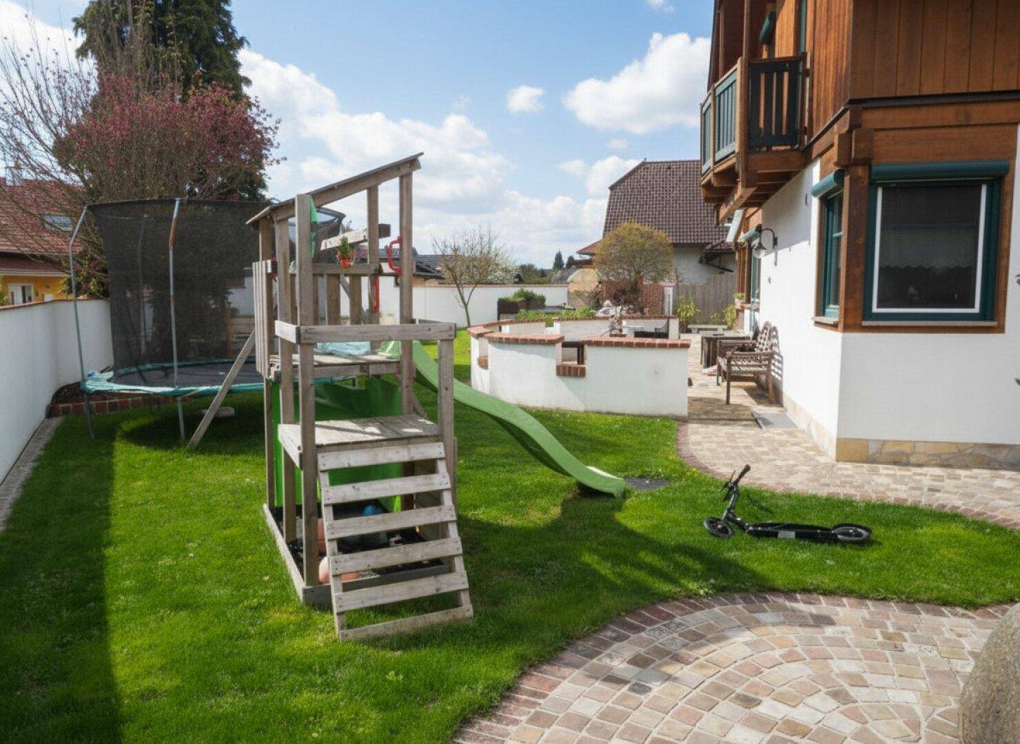 Gepflegter Garten mit Spielplatz, Trampolin, Rasenfläche und gemauerter Grillstelle neben dem Haus.