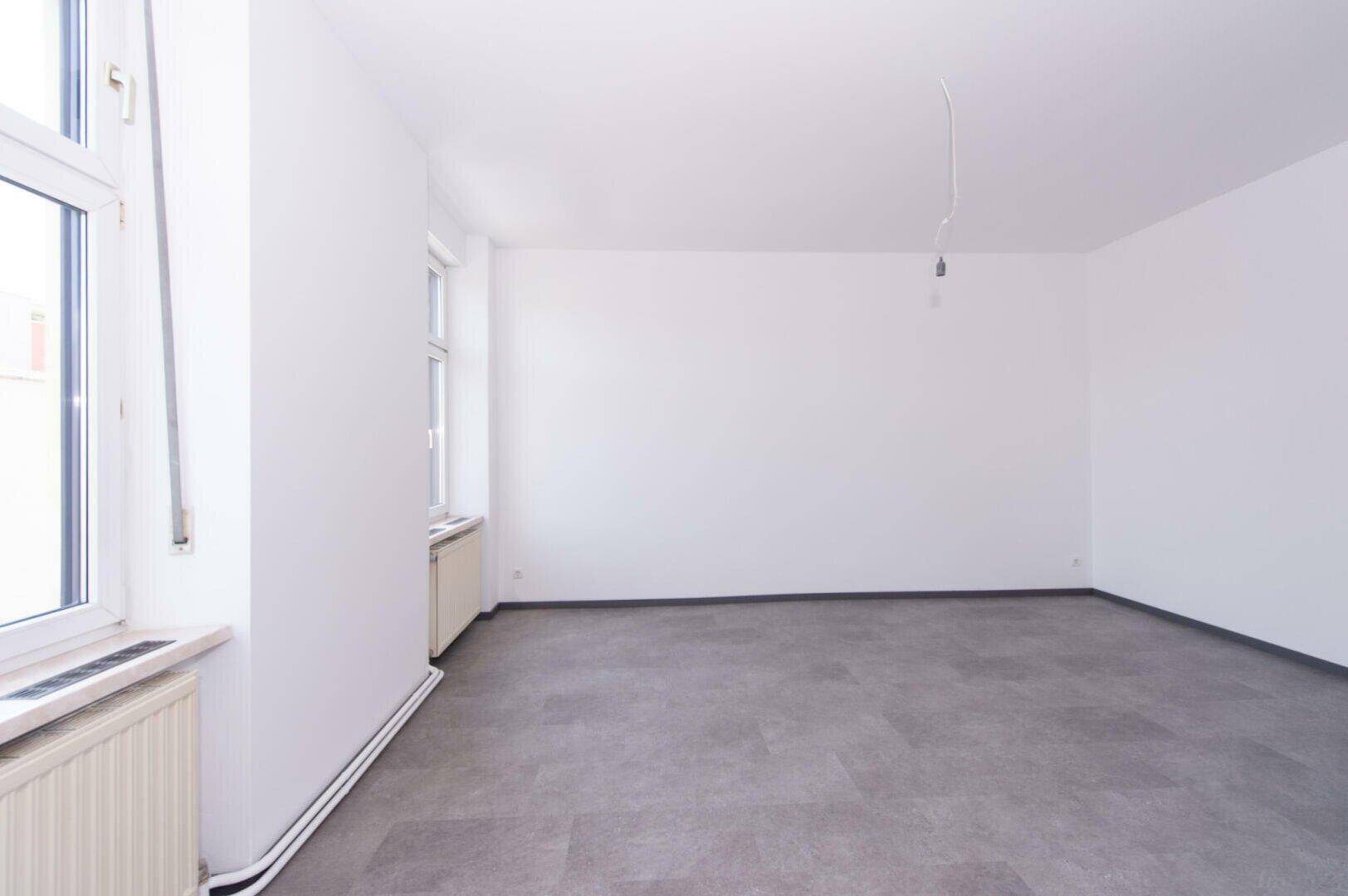 Heller, leerer Raum mit weißen Wänden, grauem Boden und zwei Fenstern mit Heizkörpern.